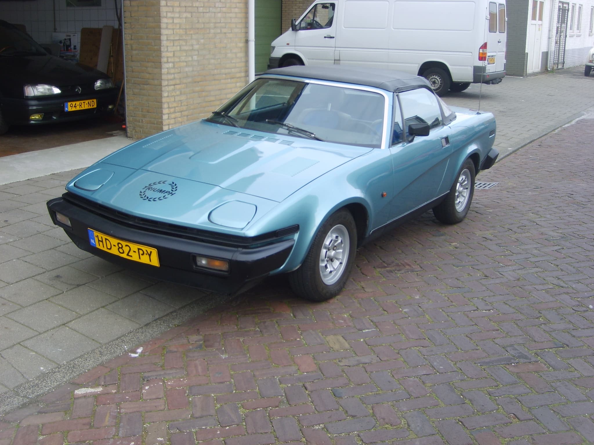 A light blue convertible with its hood up, parked in a town street.