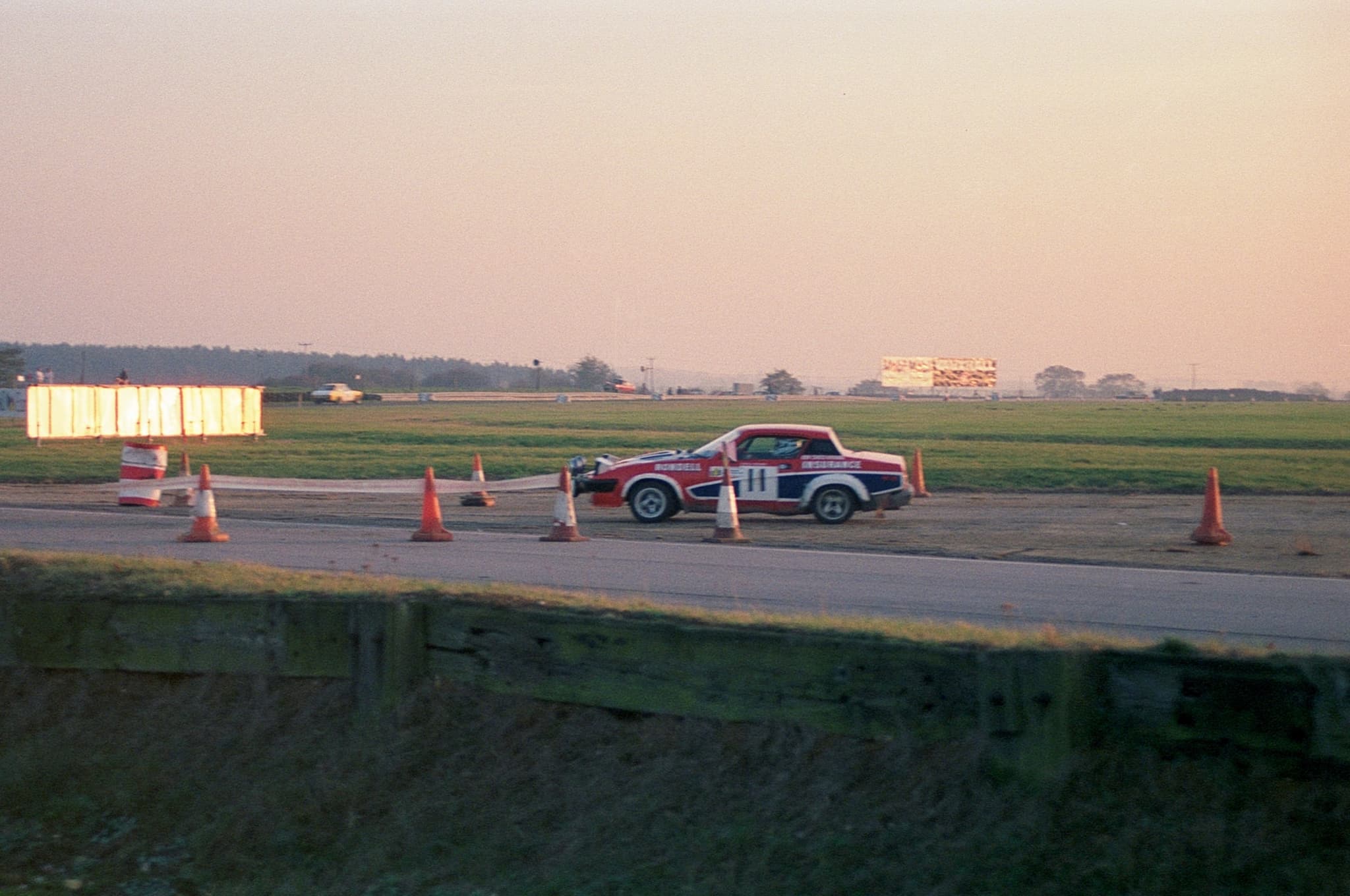 A red, white and blue rally coupé numbered 11 rounding cones on an airfield stage in low evening light. Opens a larger view