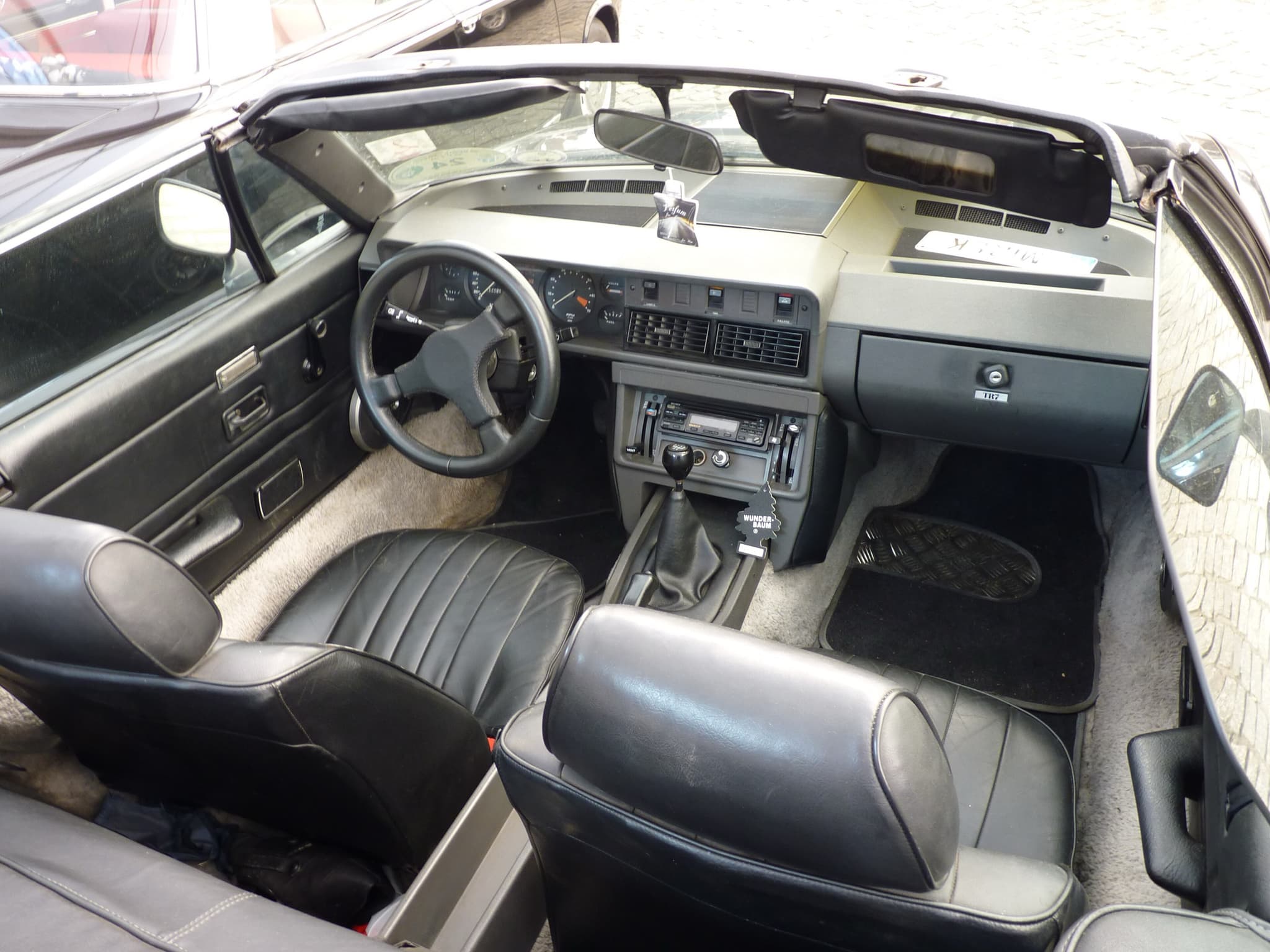 Looking down into a convertible's cockpit: dashboard, steering wheel, gear lever and black seats. Opens a larger view