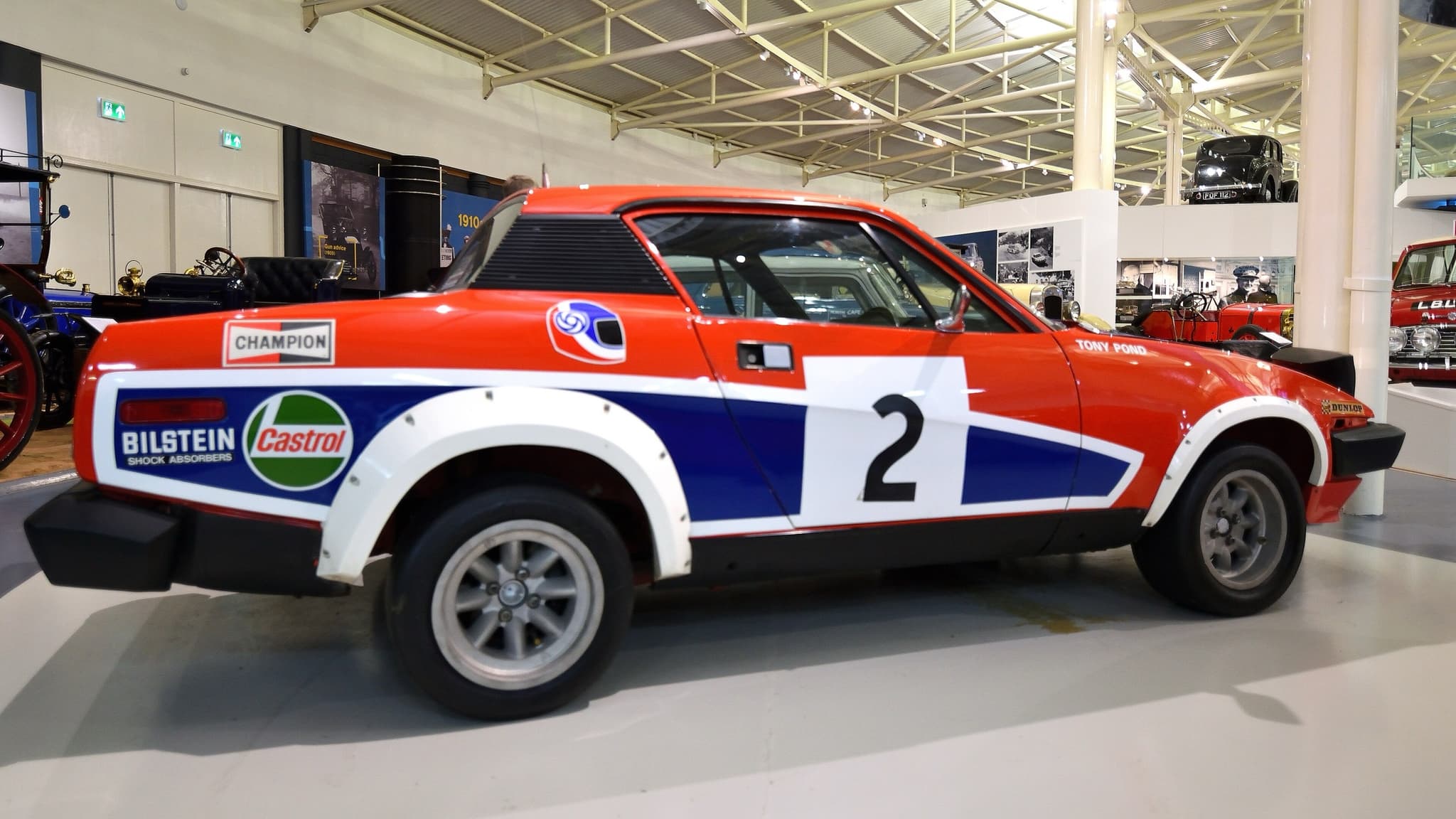 A red, white and blue rally coupé with flared arches and the number 2, seen side-on in a museum hall.