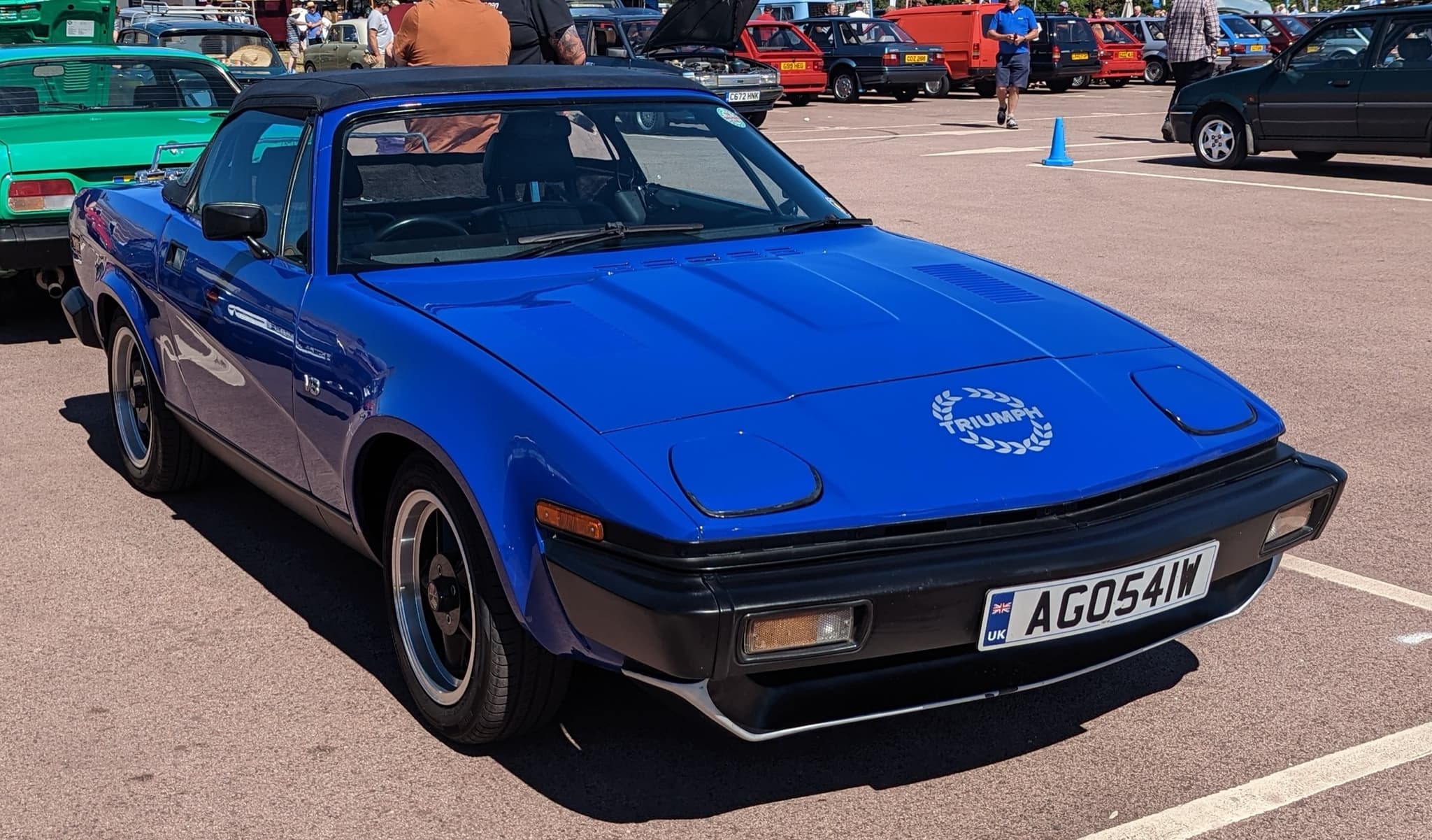 A blue convertible with its black hood up and a laurel-wreath decal, in a show car park. Opens a larger view