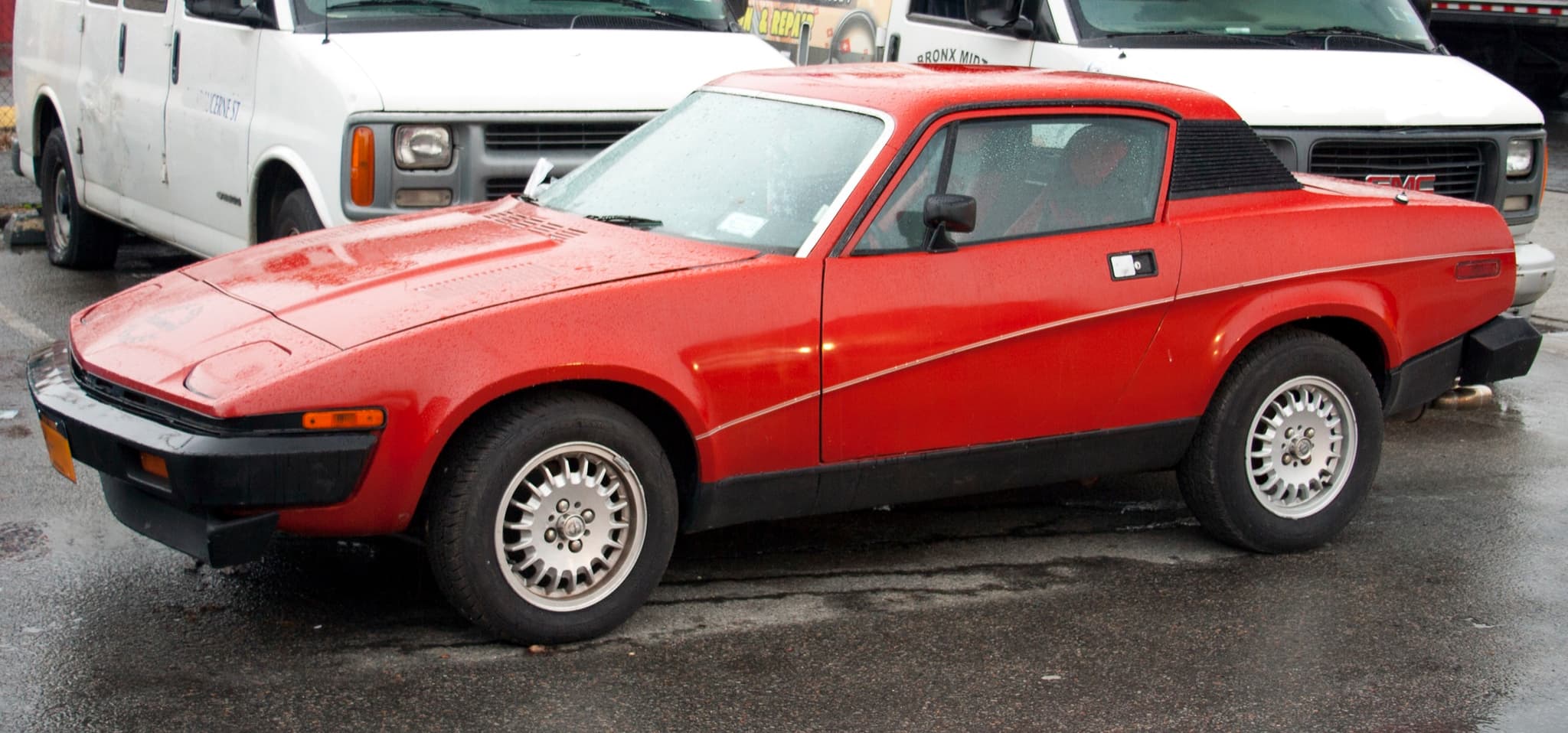 A red coupé with long black bumpers and side-marker lamps on a wet city street, vans behind.