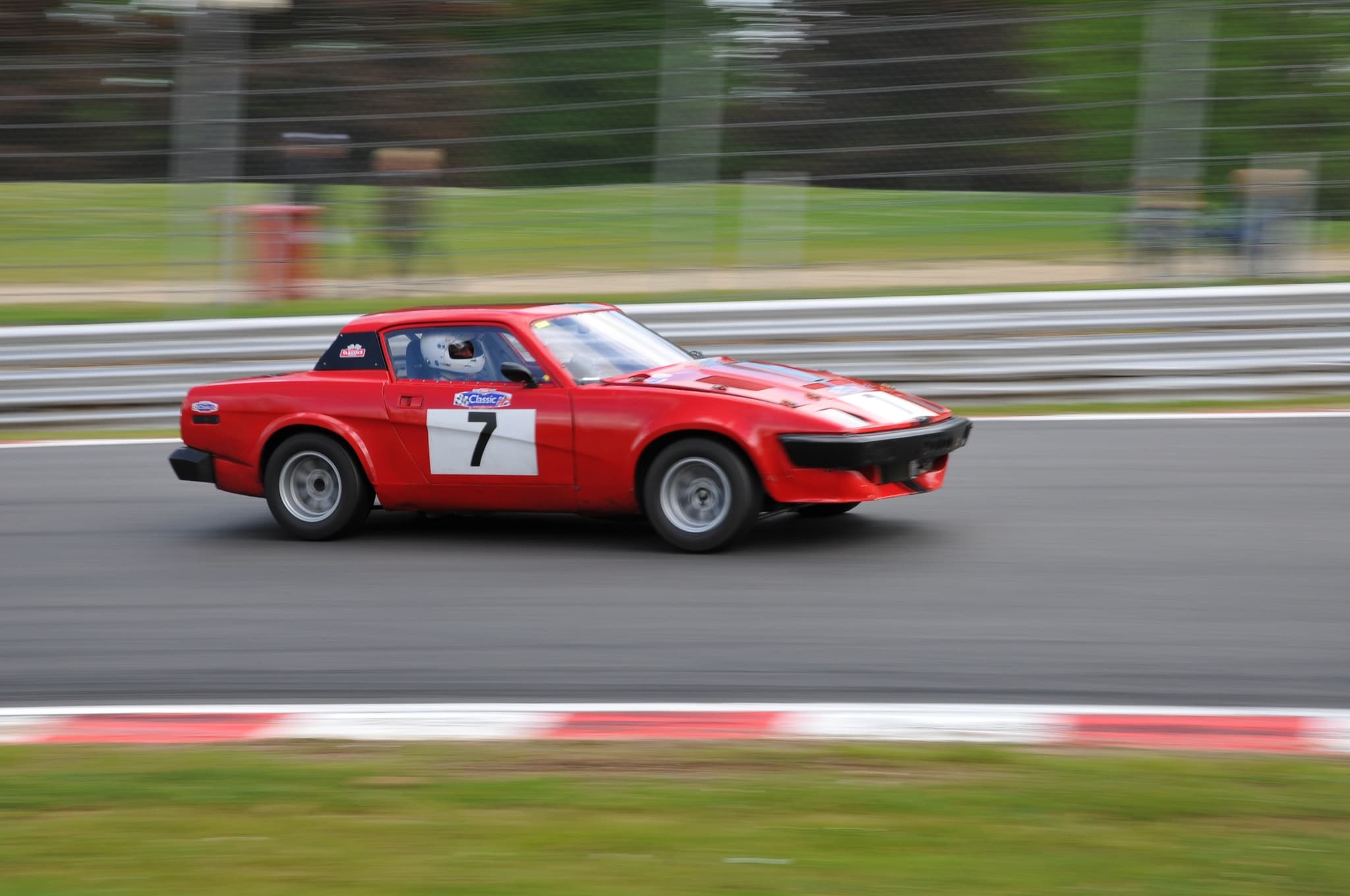 A red coupé race car numbered 7 at speed past circuit barriers, the background blurred. Opens a larger view