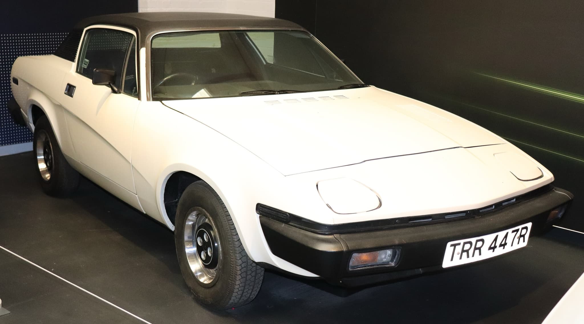 A white wedge-shaped coupé with a black vinyl roof and silver wheel trims on a dark museum stand. Opens a larger view