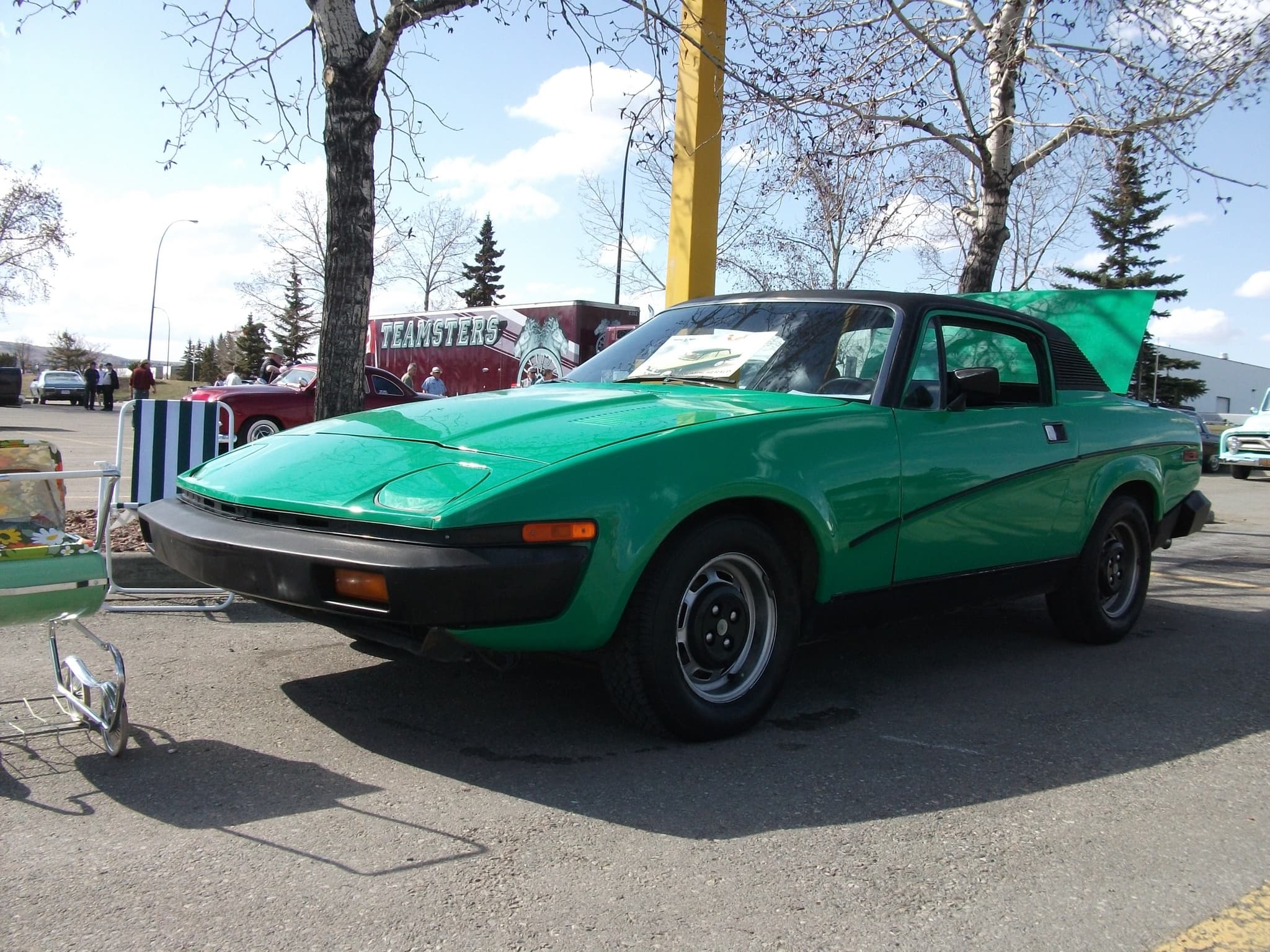 A green coupé with a black vinyl roof, long black bumpers and steel wheels at an outdoor car show, its boot open.