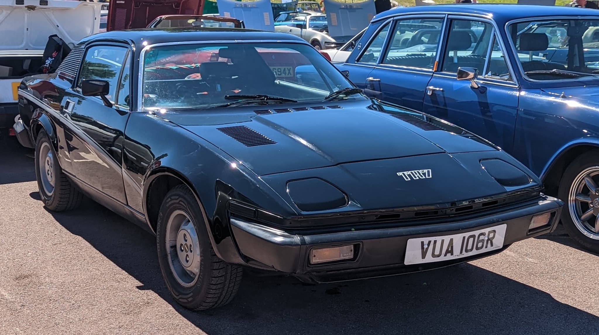 A black coupé with chrome TR7 letters on its nose and steel wheels, in bright sun.