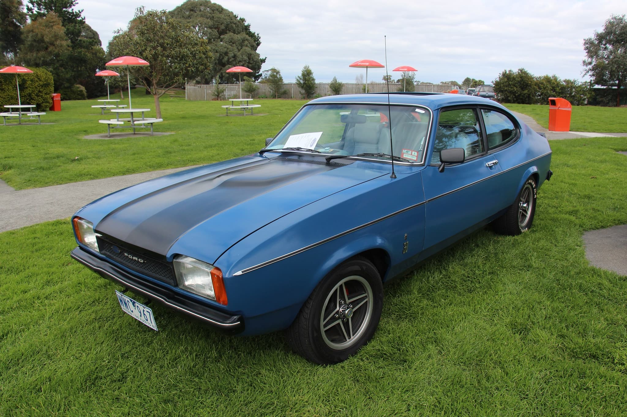 A blue coupé with a black bonnet stripe and twin headlamps, on grass.