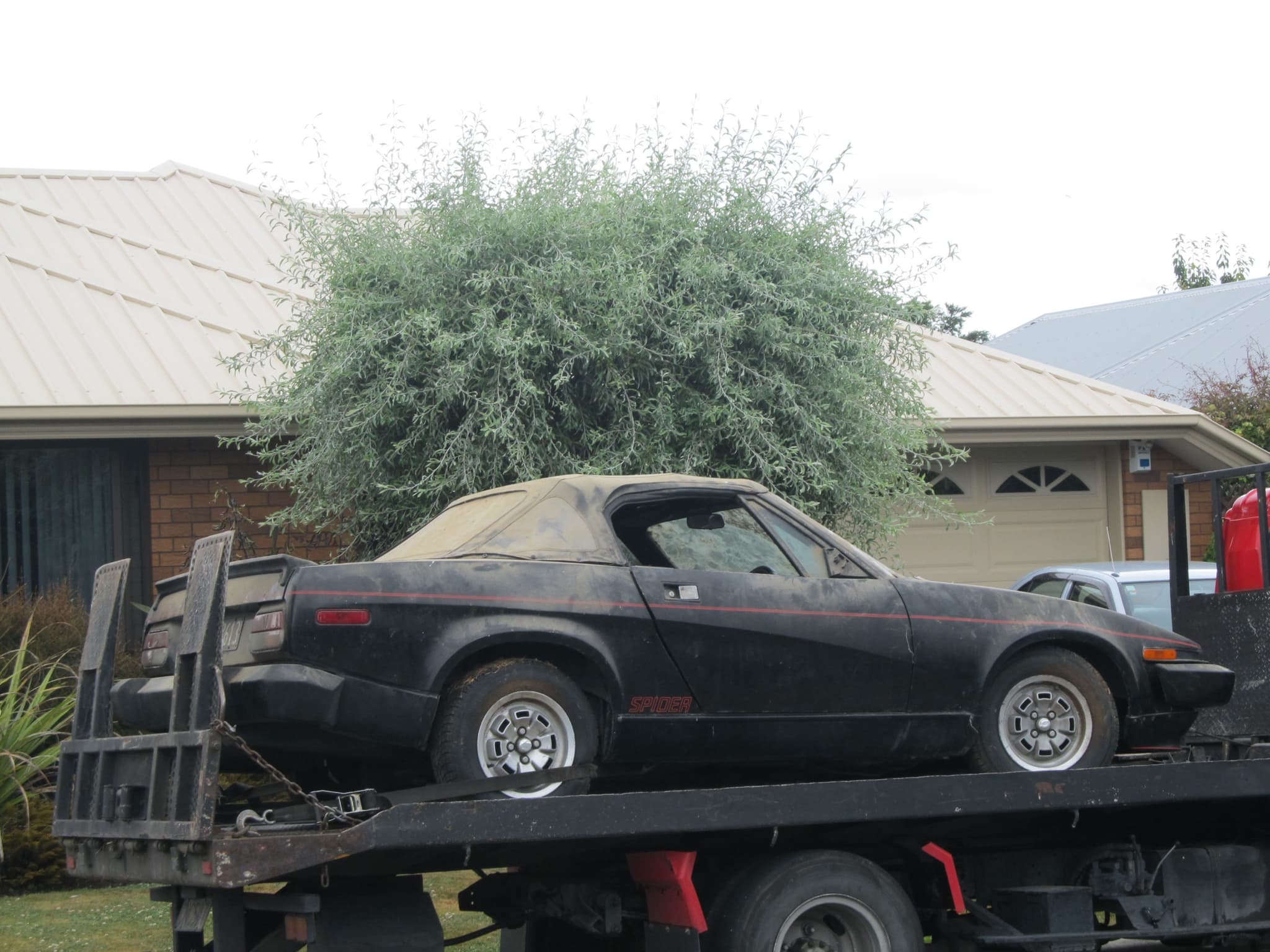 A dusty black convertible with red stripes carried side-on on the back of a flatbed truck. Opens a larger view