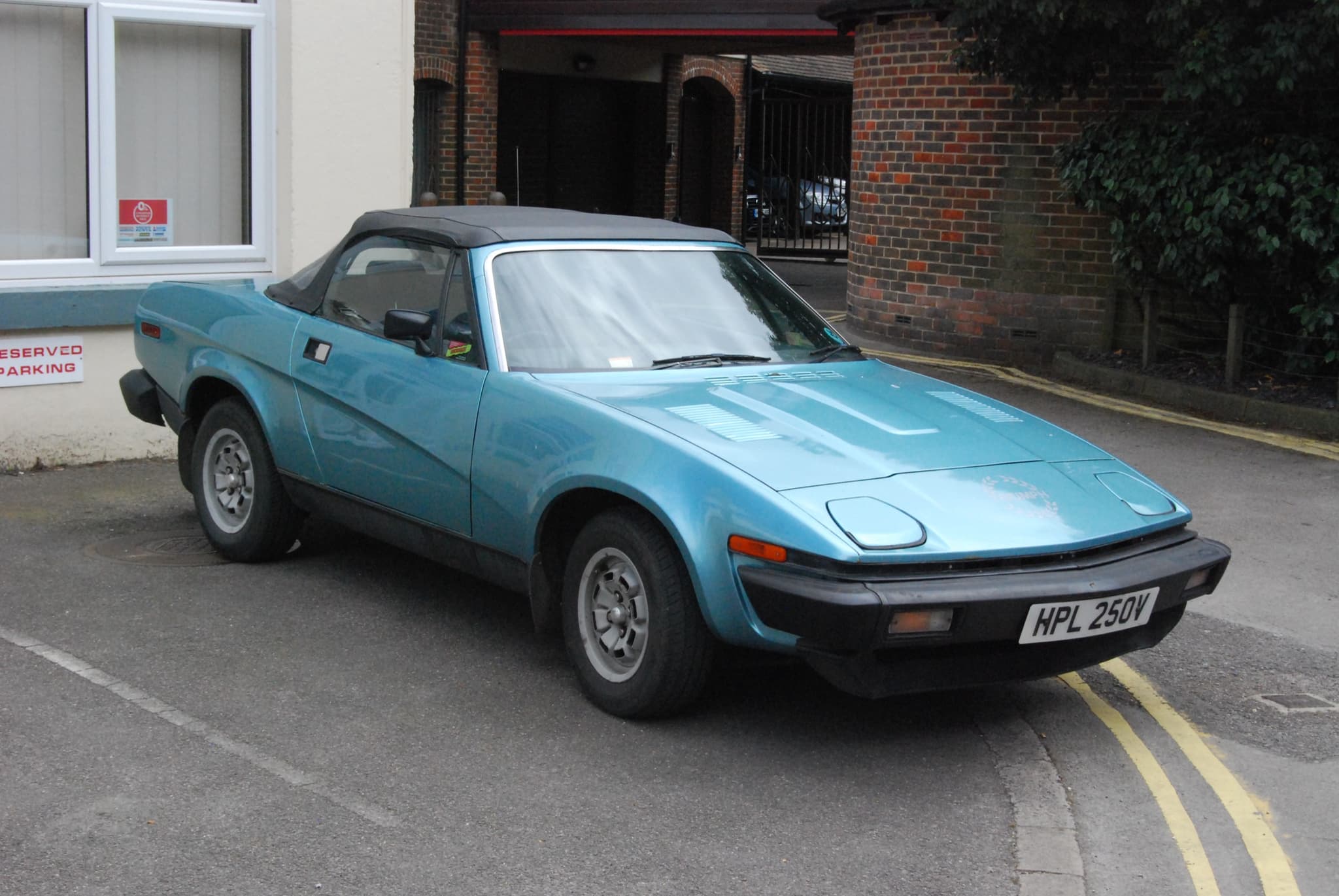 A light blue convertible with its black hood up, parked beside a white building with brick arches. Opens a larger view