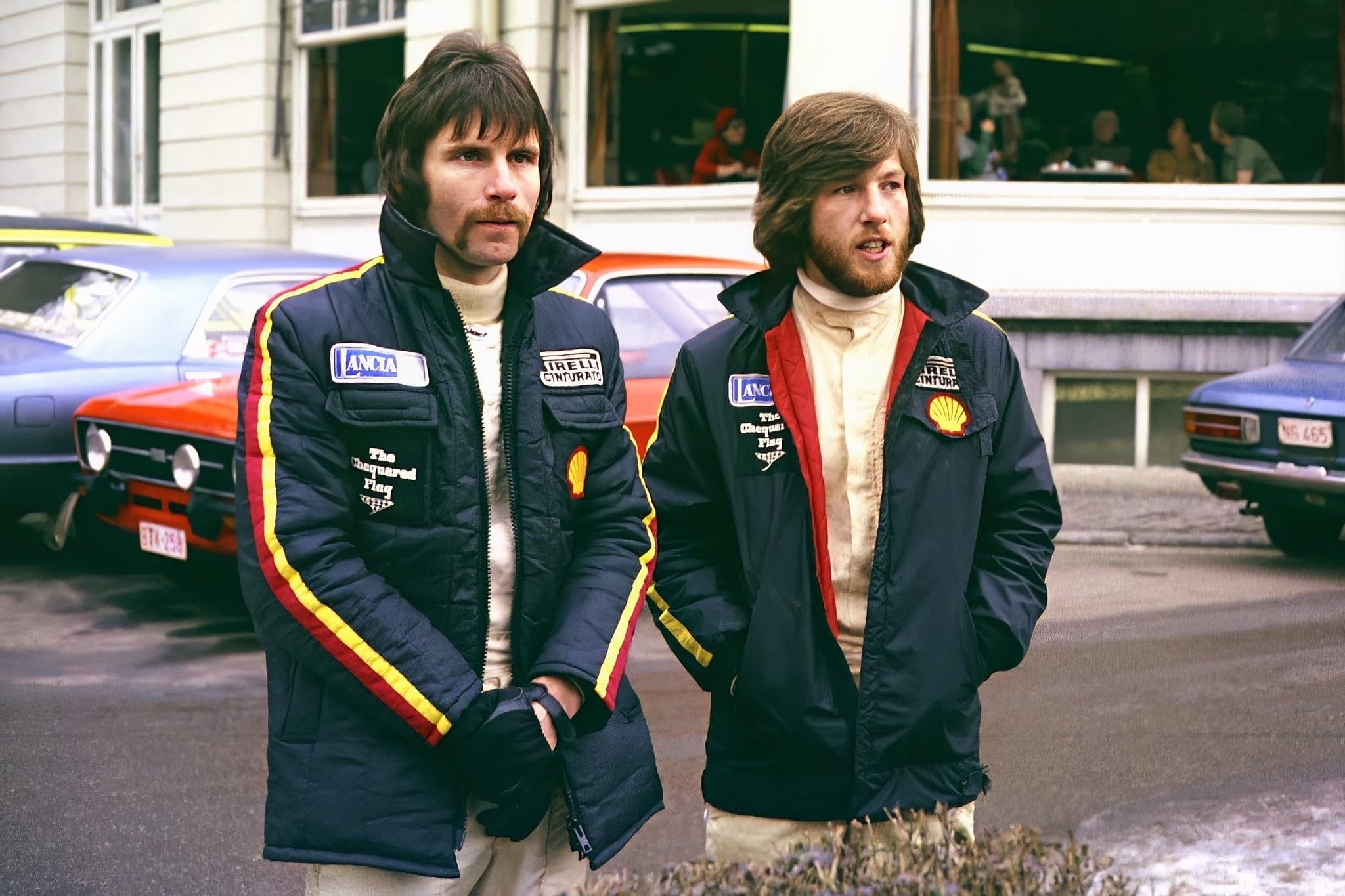 Two young men in rally team jackets stand in a street, parked cars and a café window behind them.