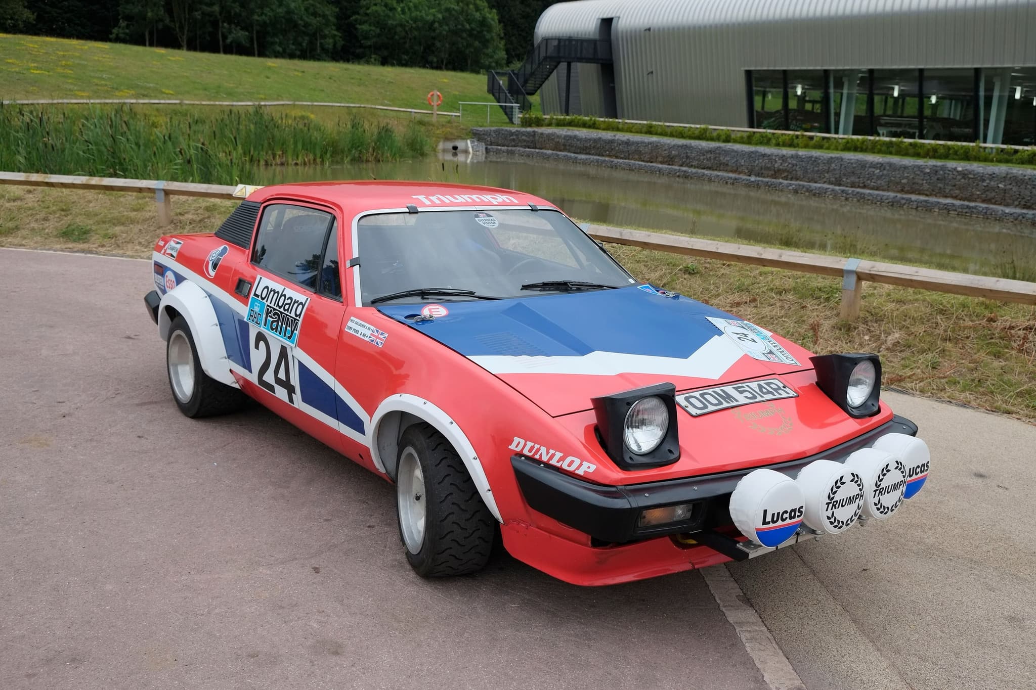 A red, white and blue rally coupé numbered 24, with four lamps on its nose, parked outside a museum building.