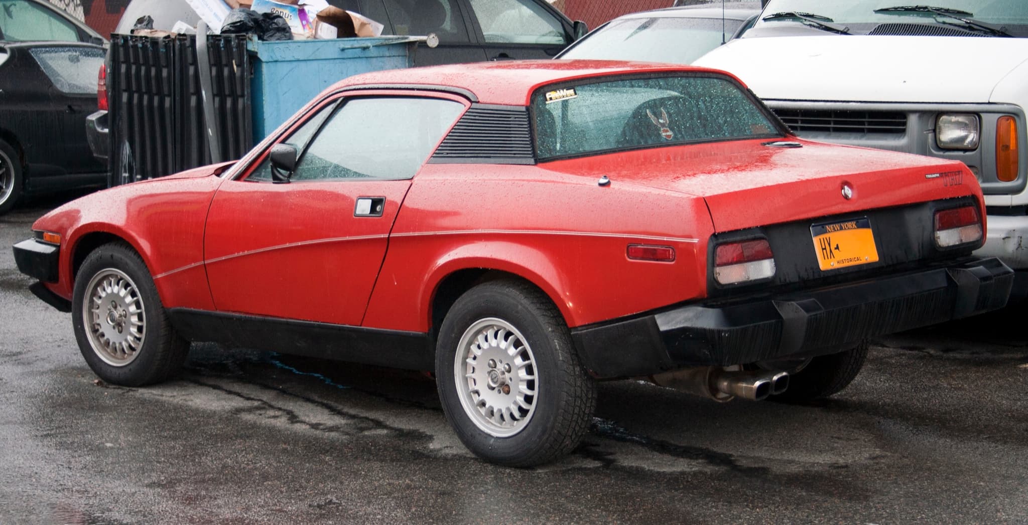 A red coupé from behind on a wet city street, its long rear bumper and side markers visible.
