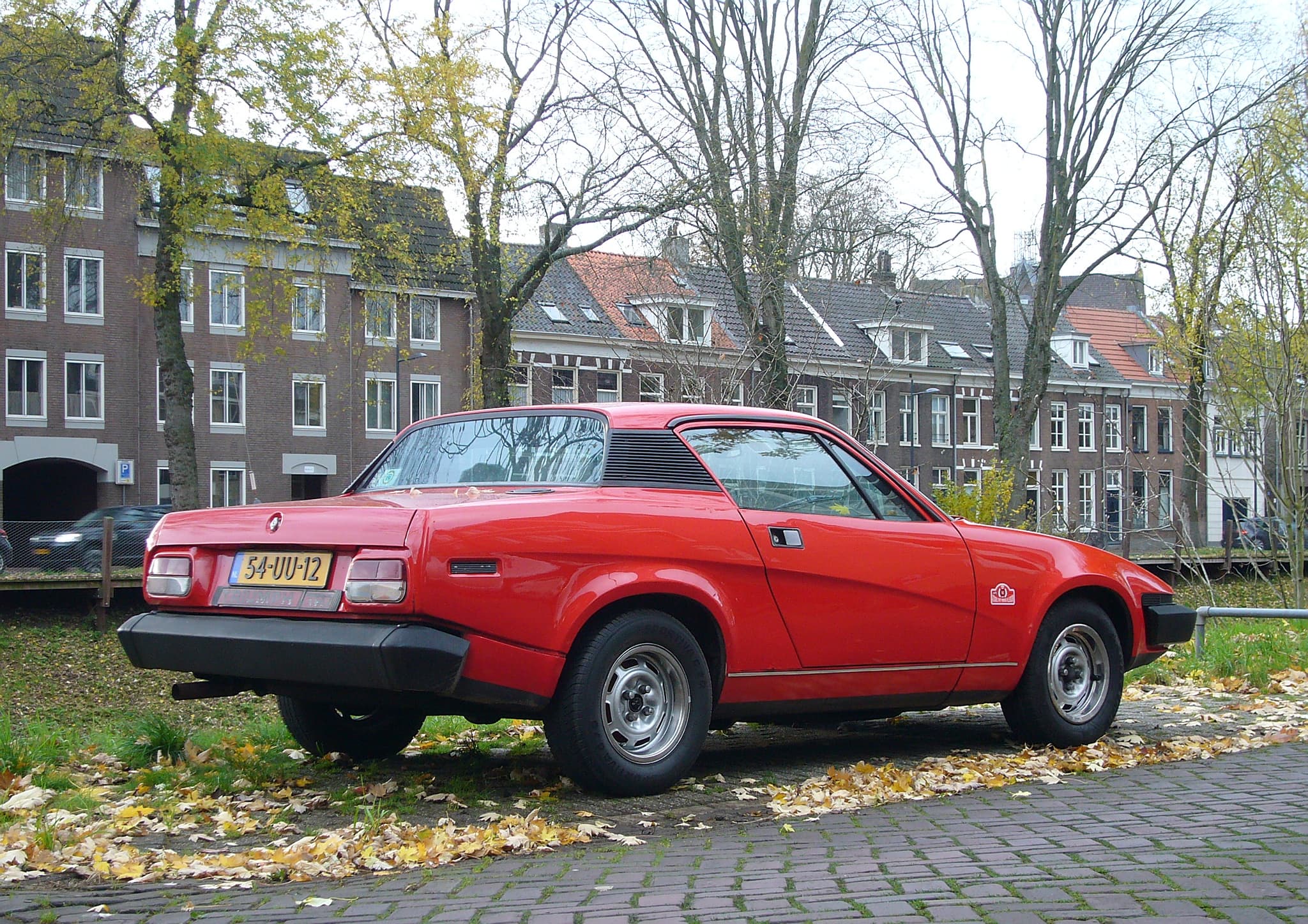 A red wedge-shaped coupé seen from behind and to one side, parked on brick paving in a street strewn with autumn leaves.