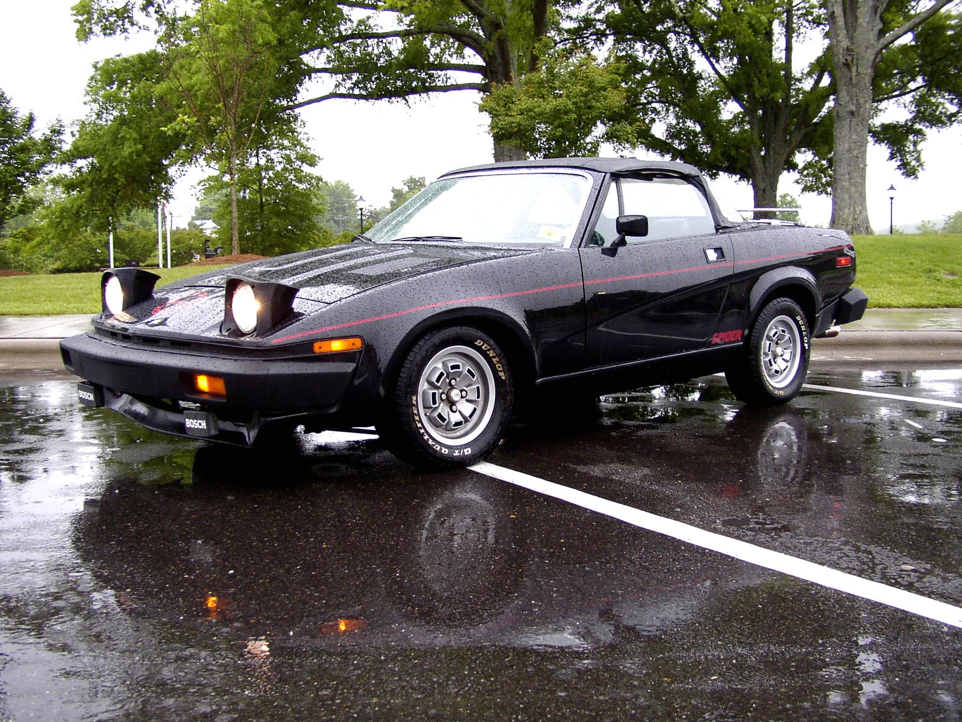 A black convertible with red side stripes and Spider badging on a wet car park. Opens a larger view