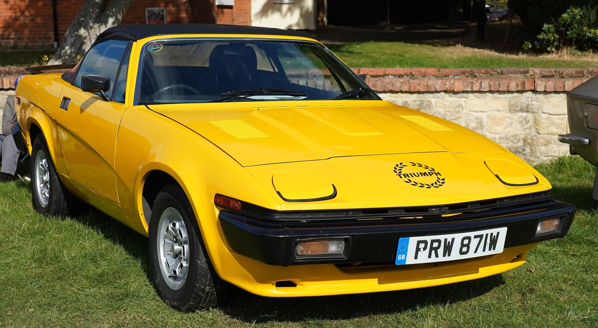 A yellow convertible with its black hood up, a laurel-wreath decal and alloy wheels, on grass in front of a brick wall.