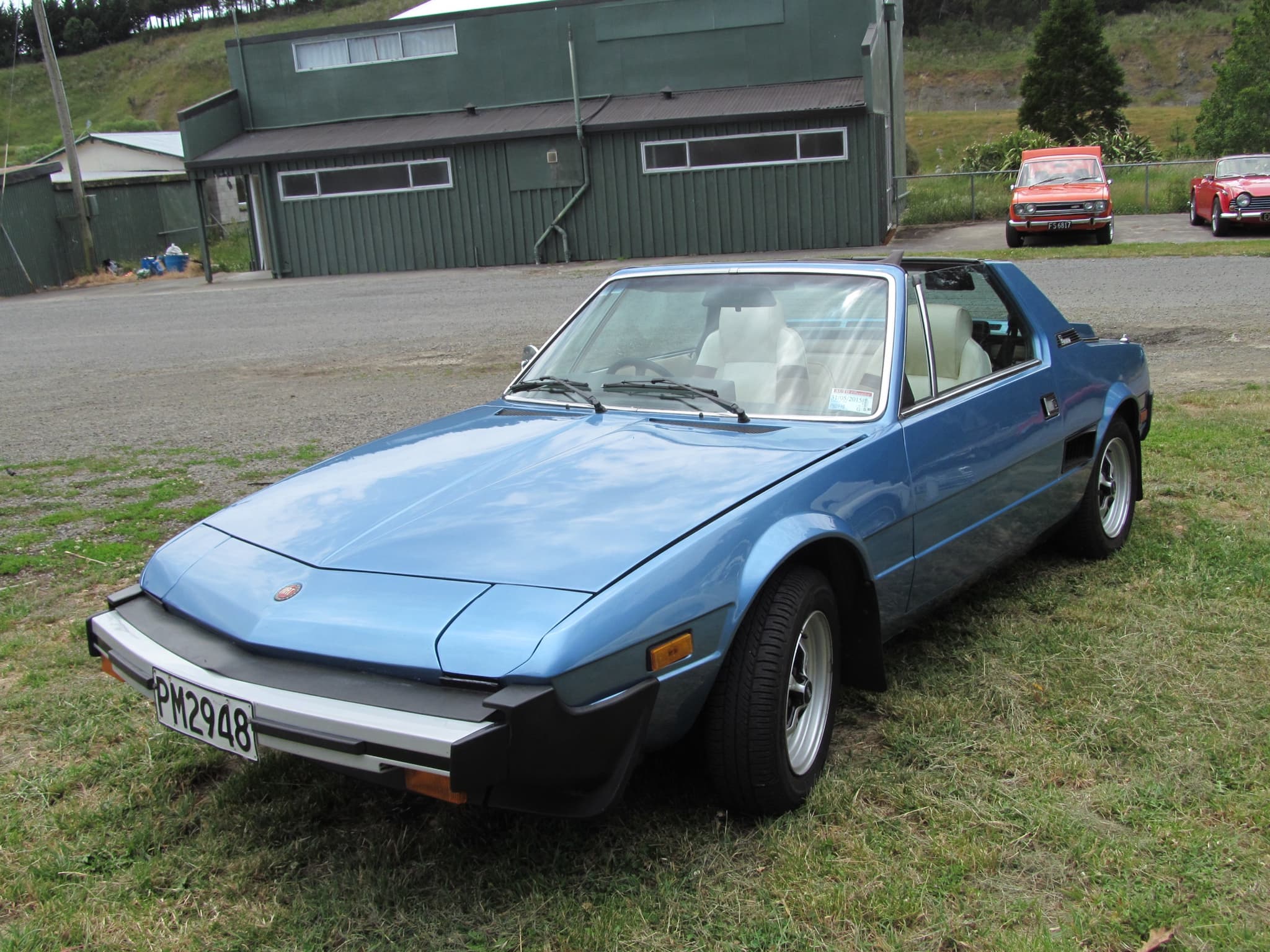 A light blue small mid-engined sports car with its roof panel off, by a farm shed.