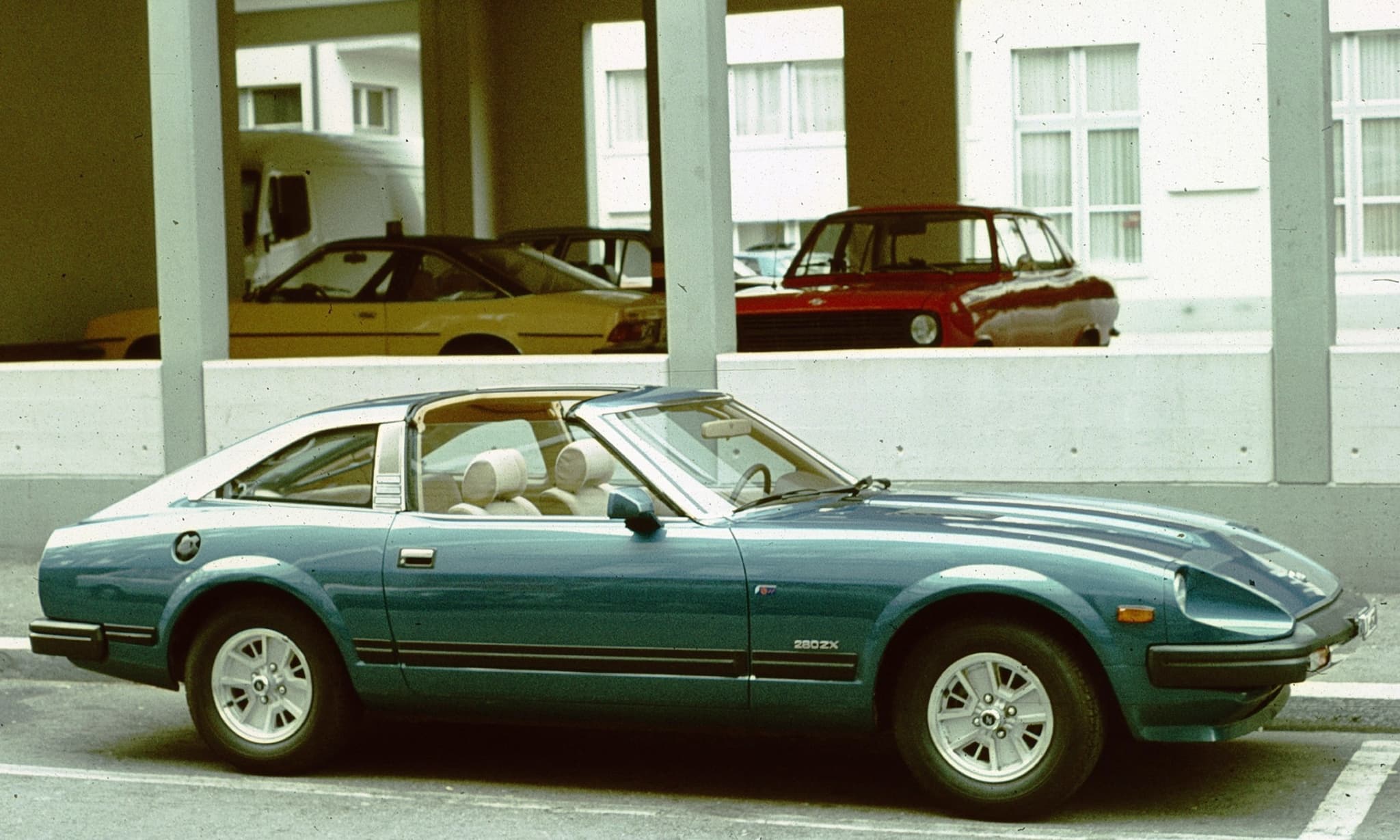 A fastback coupé with a T-bar roof and side stripes, seen in profile beneath a building in a period colour photograph.