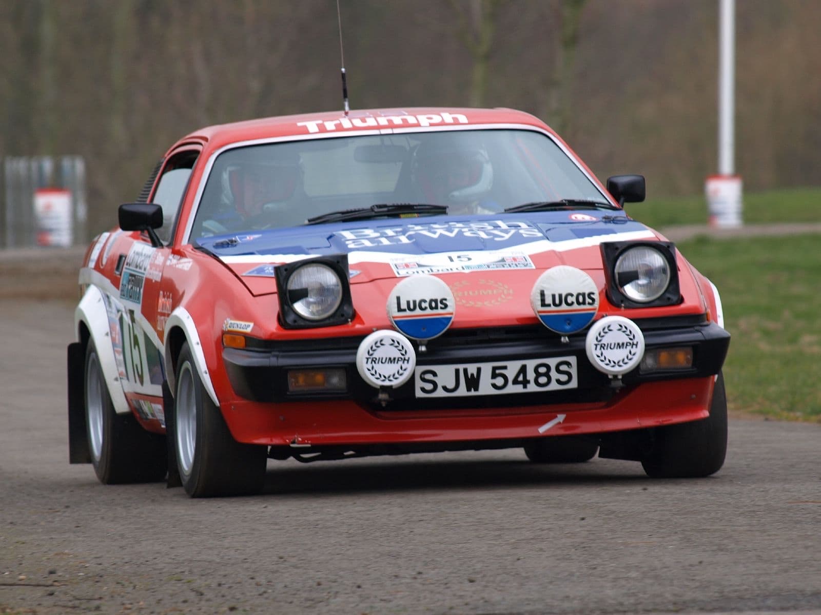 A red, white and blue rally car coming head-on along a stage, lamps covered on its nose. Opens a larger view