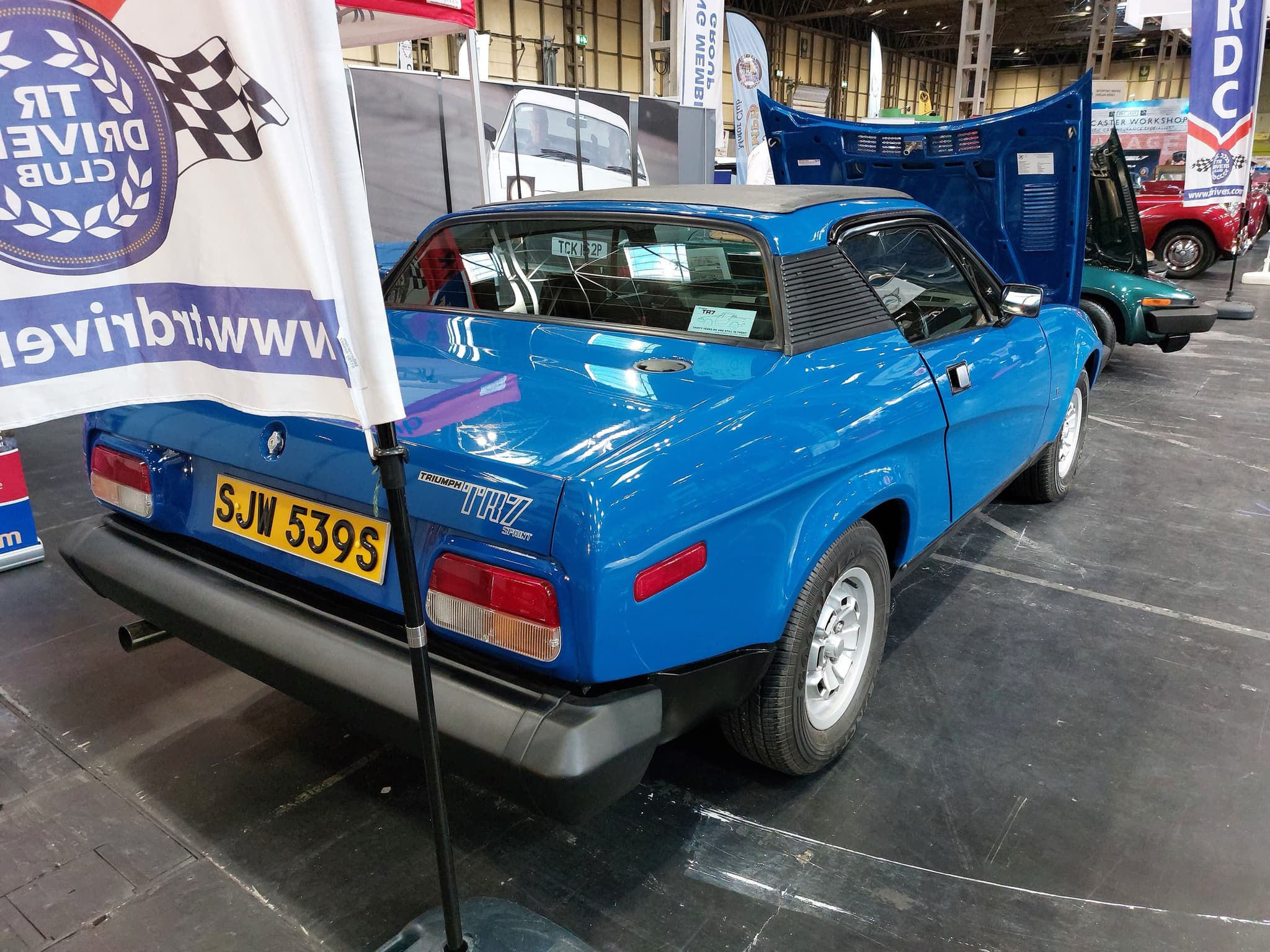 A blue coupé seen from behind on a club stand at an indoor show, with TR7 Sprint badges on its tail and a club flag beside it.