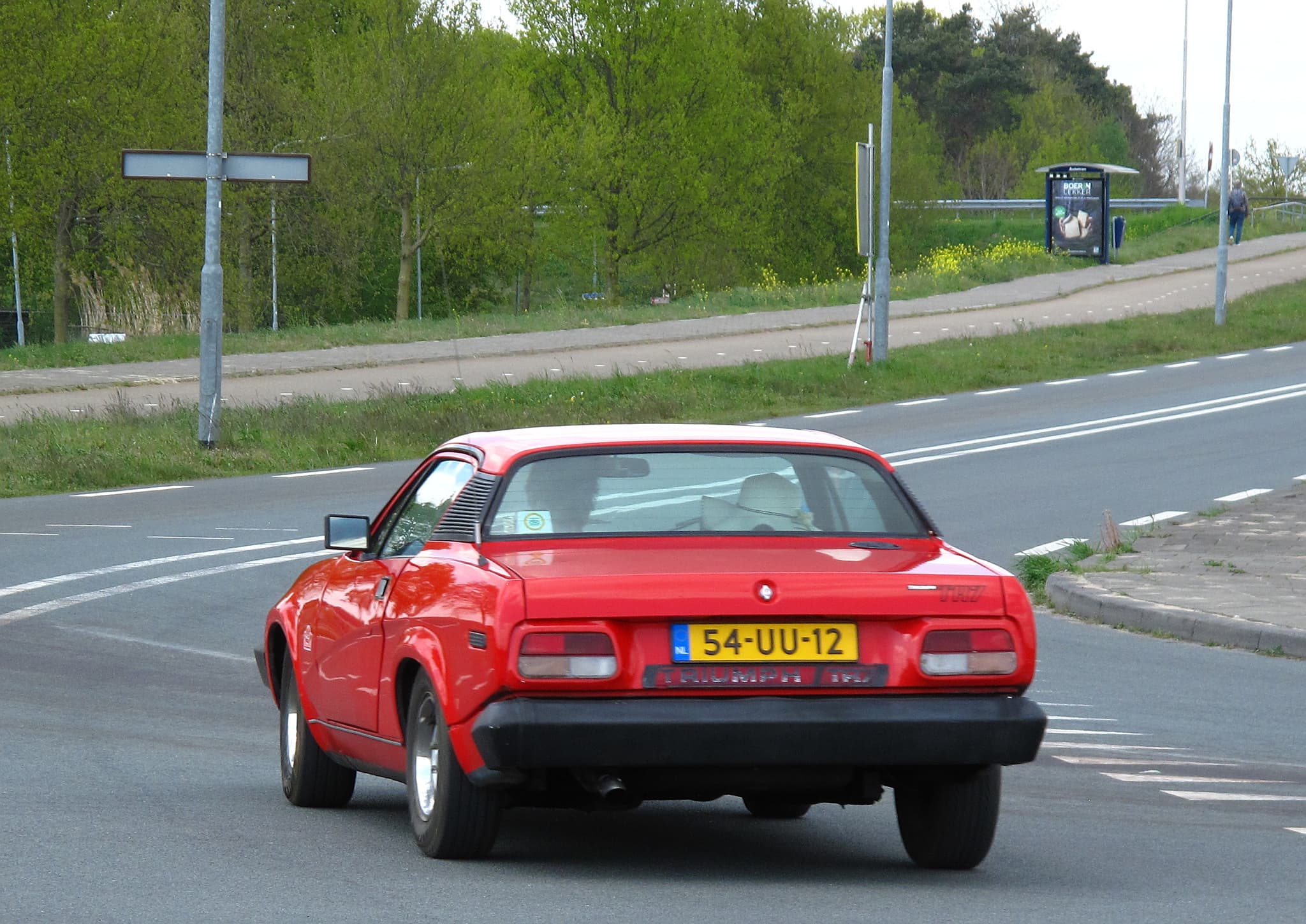 A red wedge-shaped coupé driving away along a two-lane road between grass verges and trees.