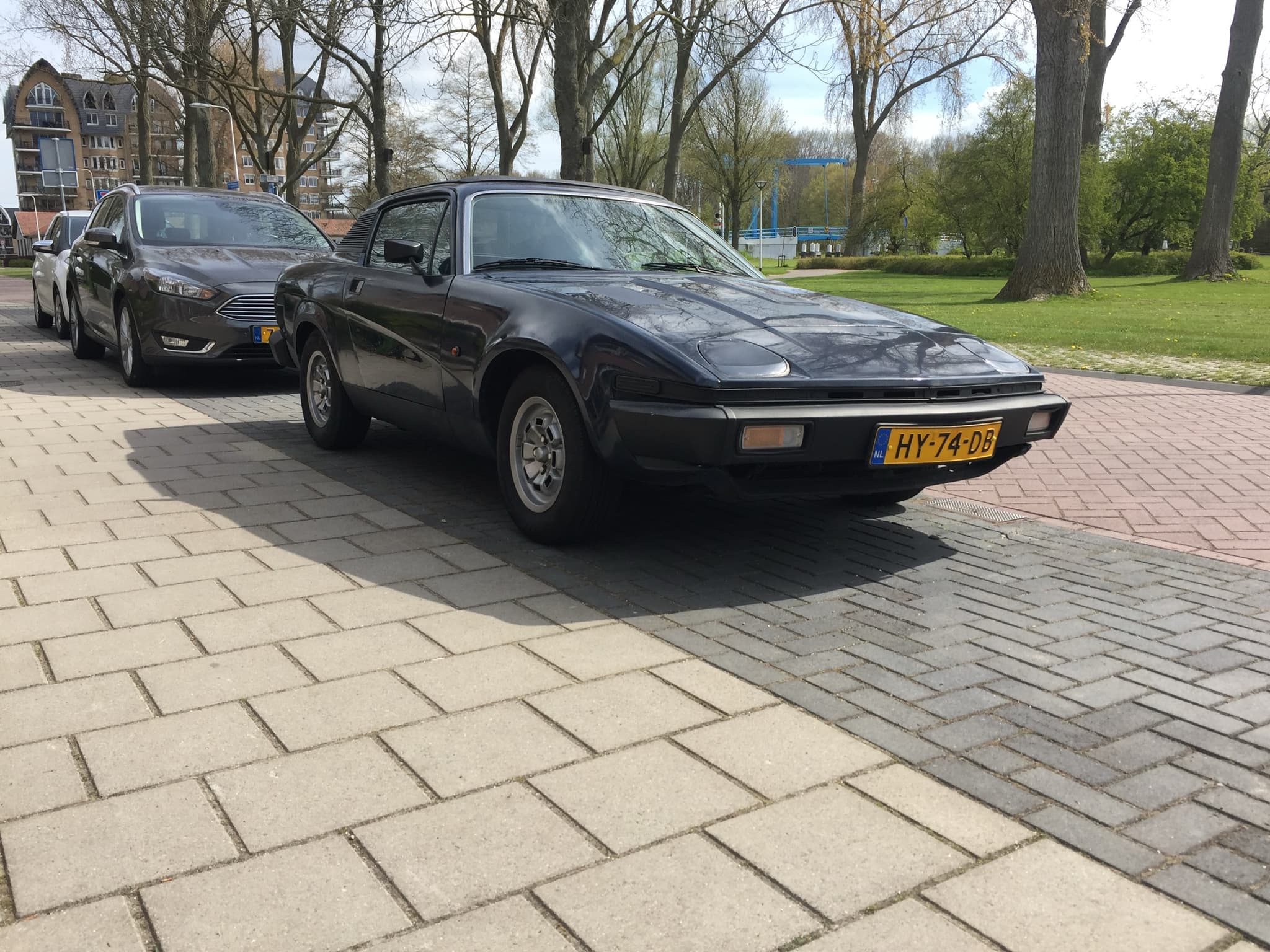 A dark blue wedge-shaped coupé parked on brick paving in a tree-lined street.