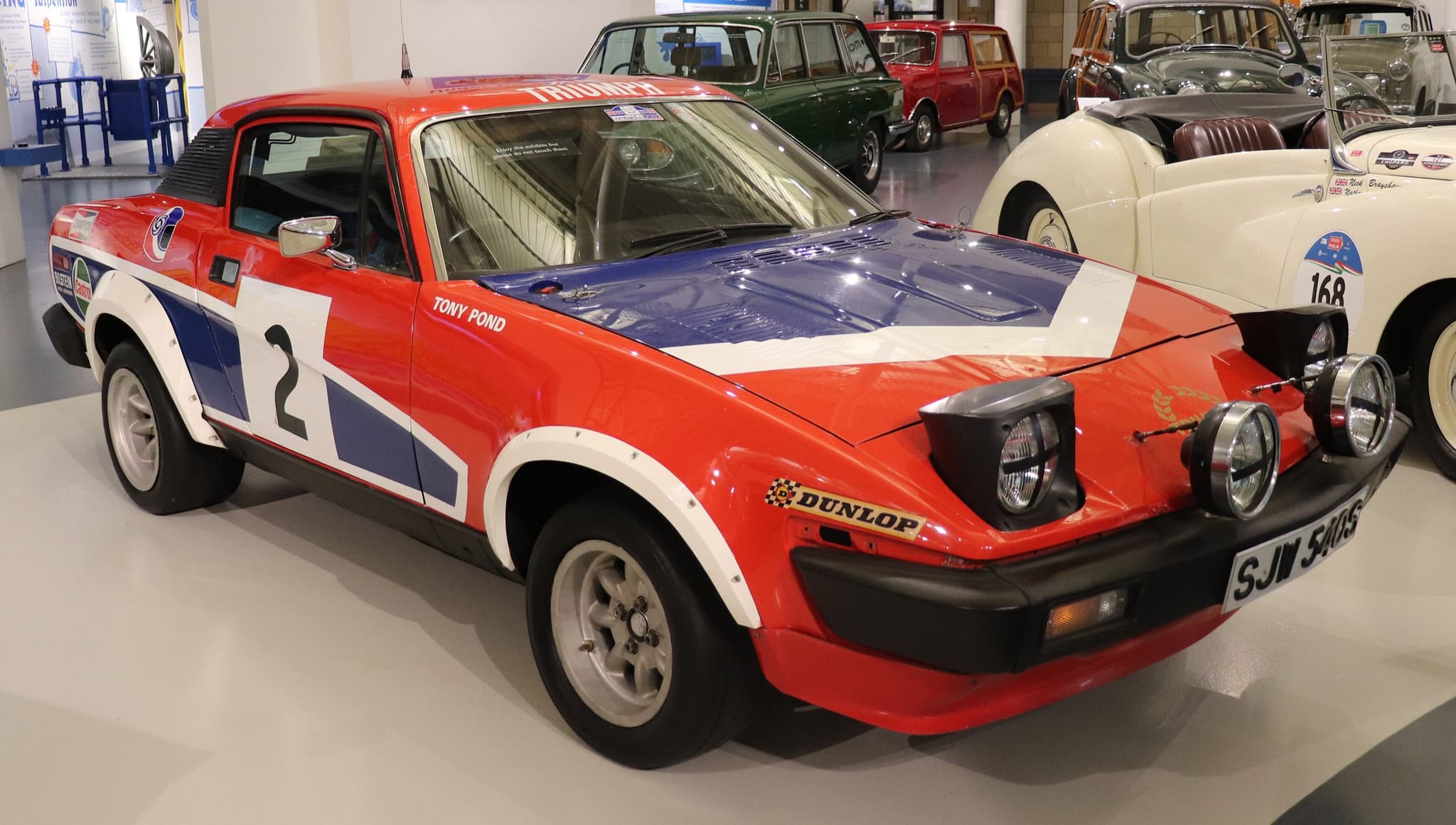 A red, white and blue rally coupé with lamp pods on its nose, numbered 2, in a museum hall.