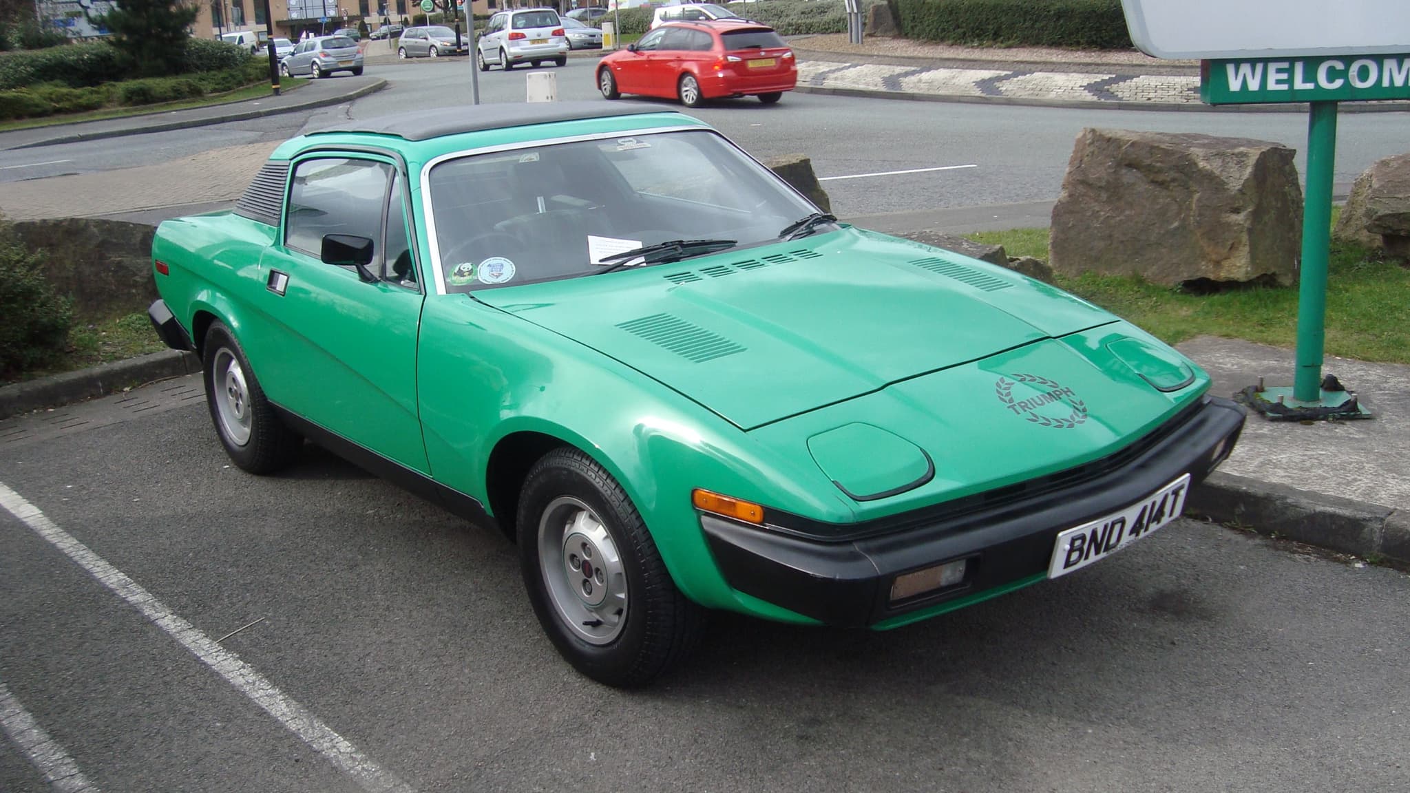 A bright green coupé with a laurel-wreath Triumph decal on its nose, parked beside a roundabout.