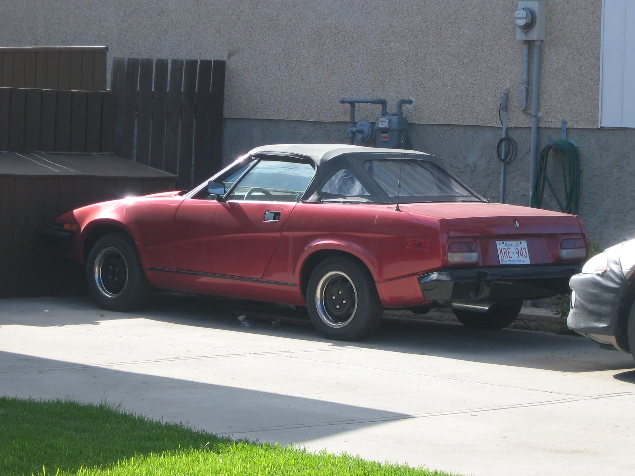 A red convertible with its hood up and long black bumpers, side-on beside a building.
