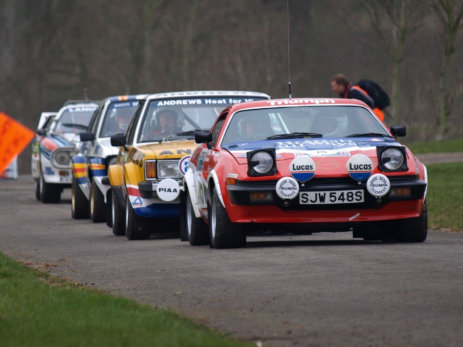 A red, white and blue rally car leading a queue of older rally cars along a tarmac stage.