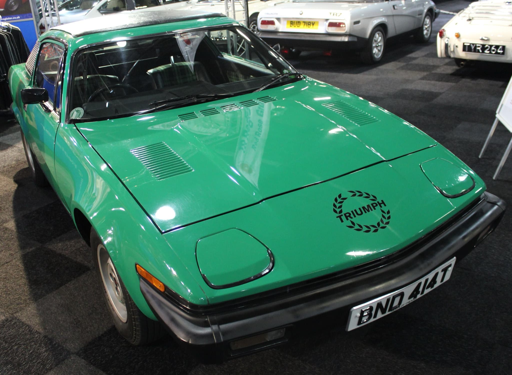 A green coupé with a laurel-wreath Triumph decal on its nose, seen close from above on a show floor.