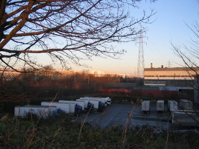 Industrial units and parked lorry trailers at dusk, seen through bare trees.