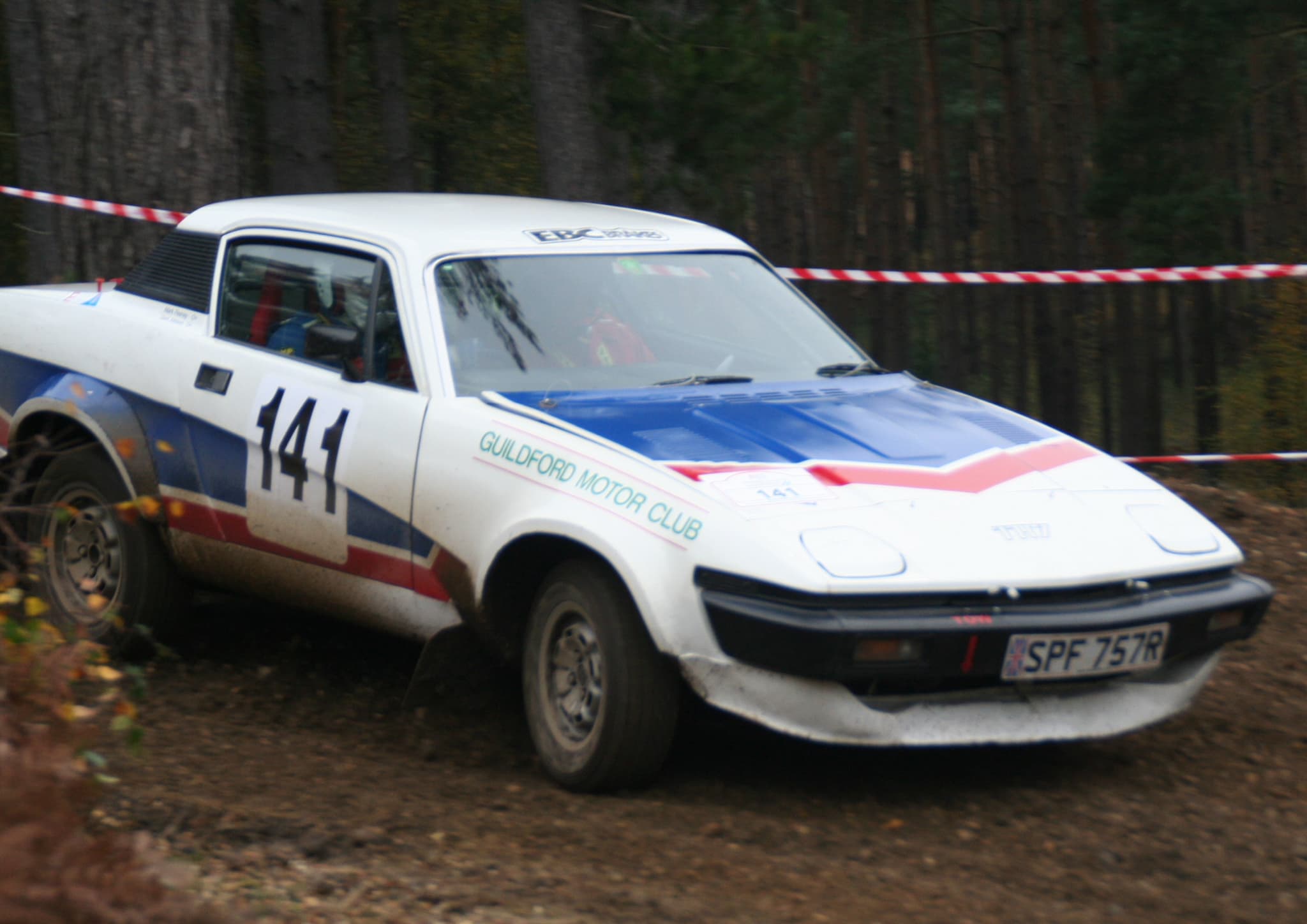A white rally coupé with a red, white and blue bonnet, numbered 141, sliding through mud on a forest stage.
