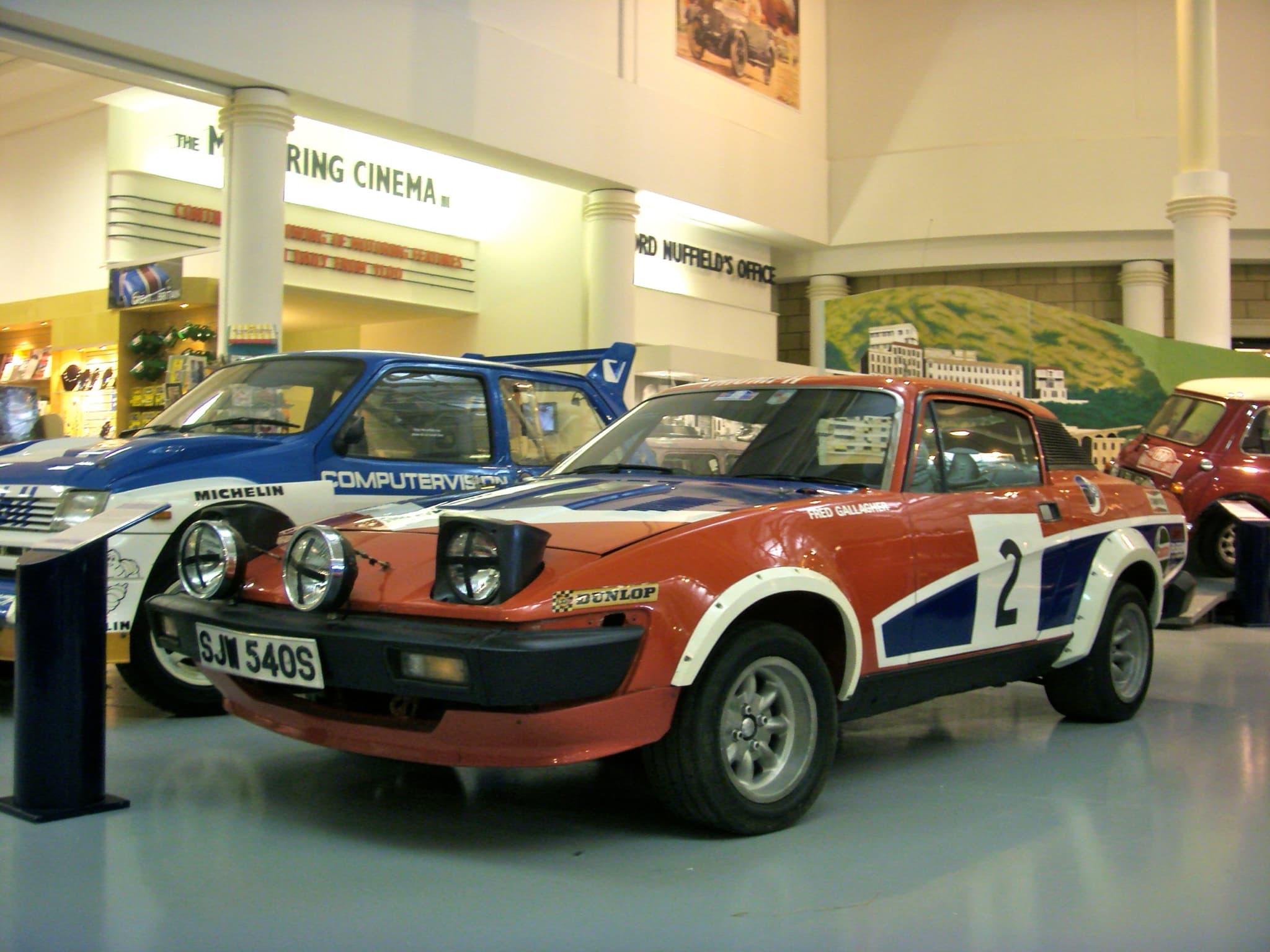A red, white and blue rally car numbered 2 in a museum hall, a white rally hatchback behind.
