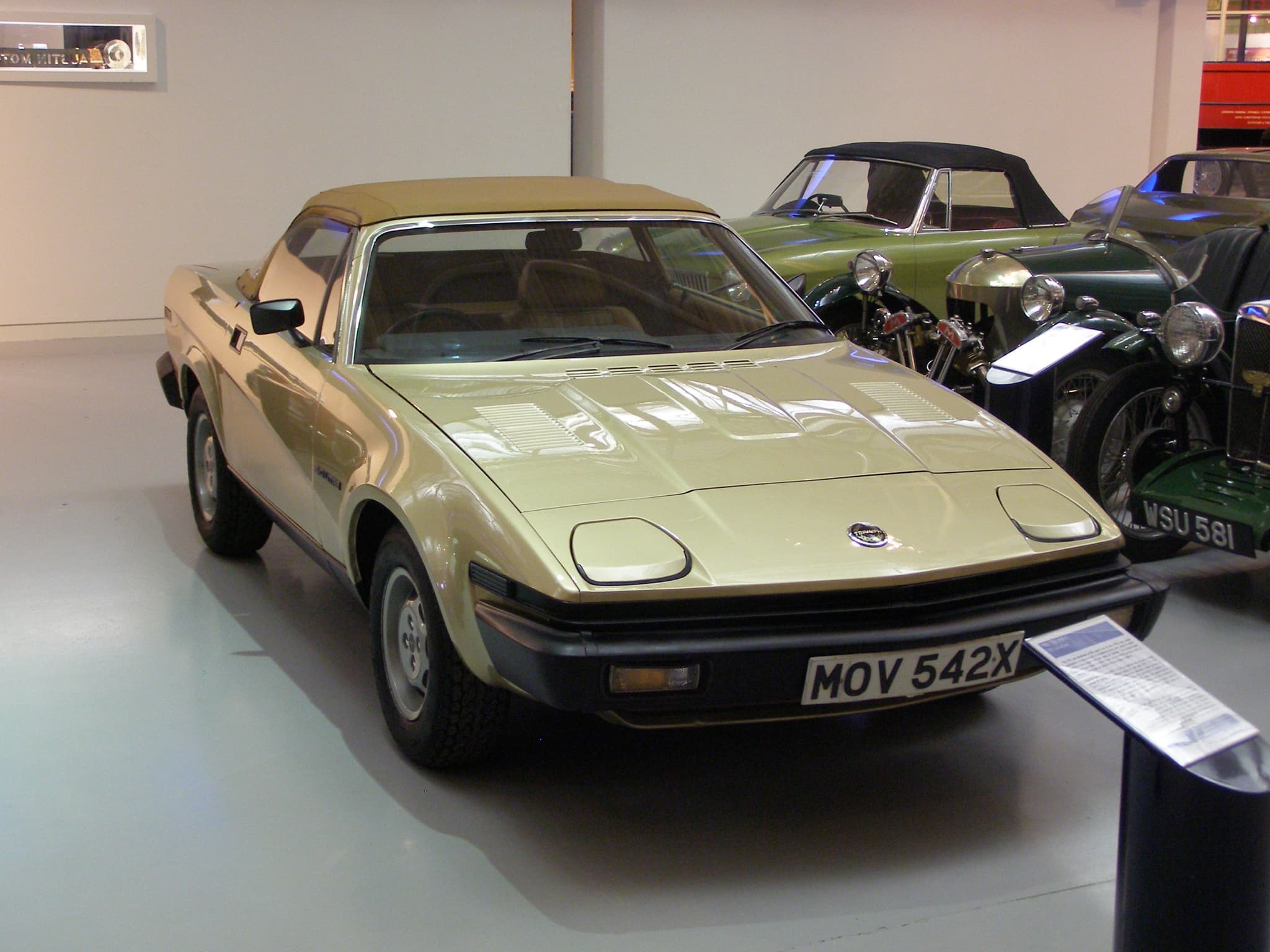 A champagne convertible with its hood up beside an information board in a museum.