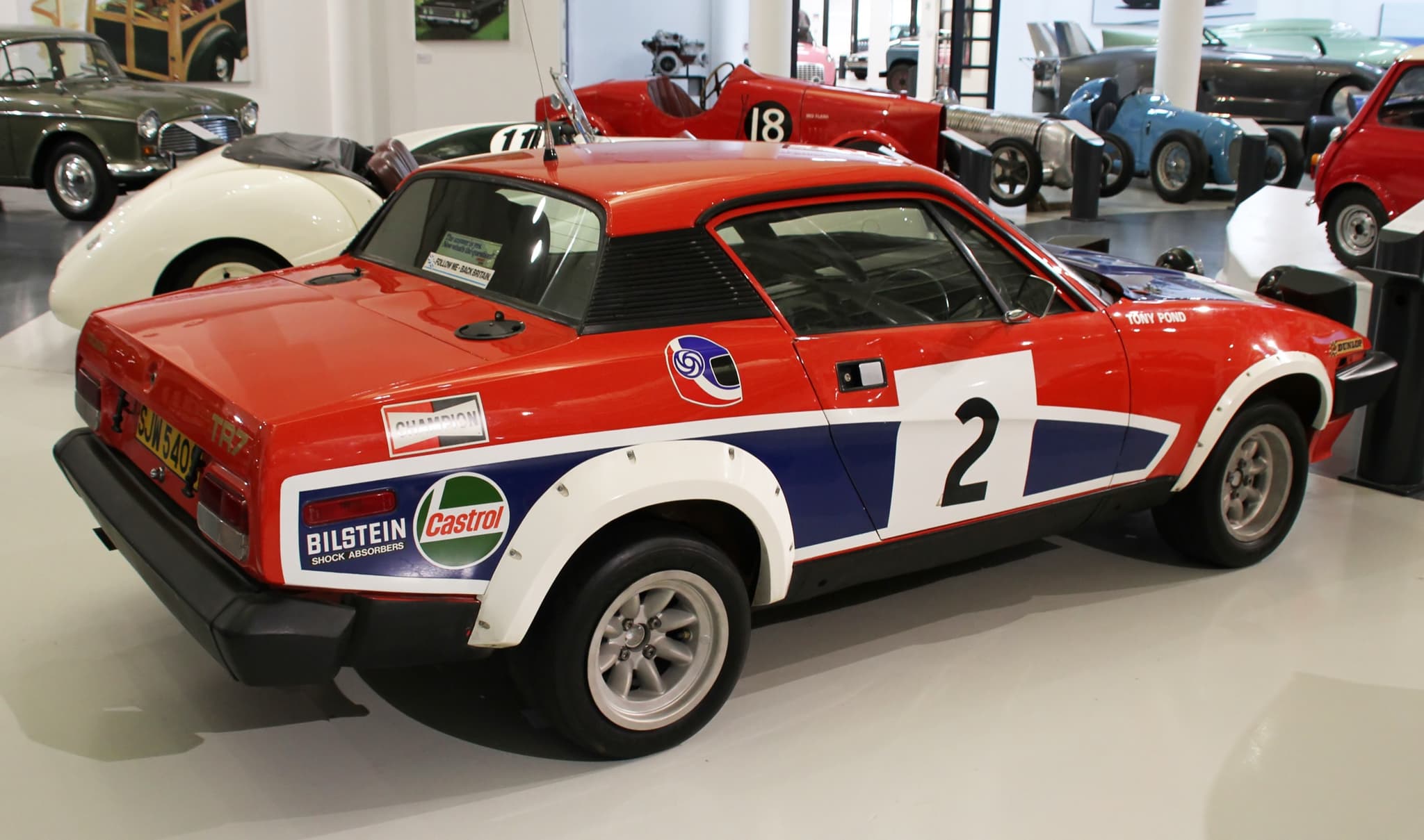 A red, white and blue rally car numbered 2 seen side-on, a driver's name lettered by the window, in a museum.
