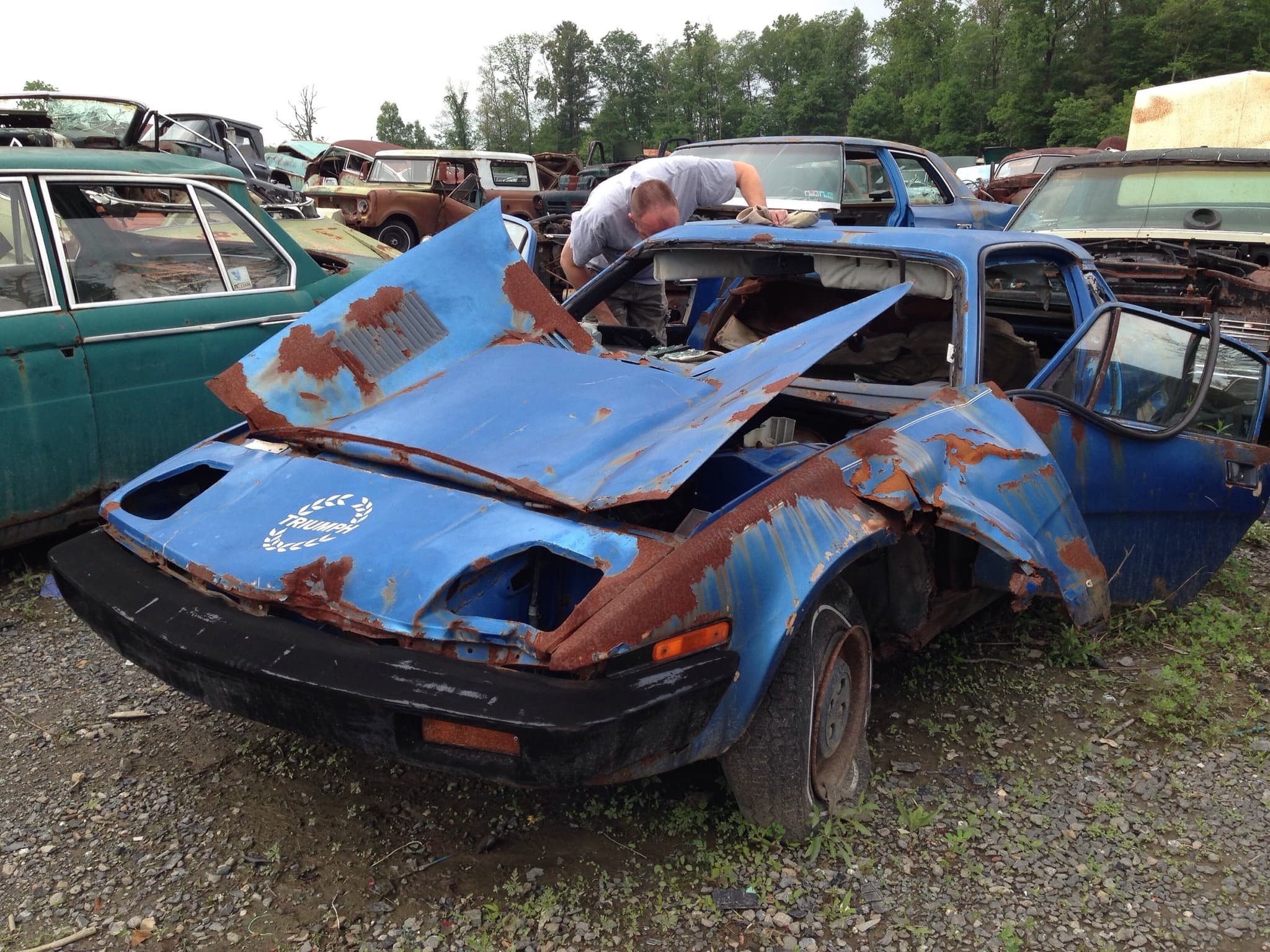 A blue coupé rusted through and crumpled at the front, standing among wrecks in a scrapyard.