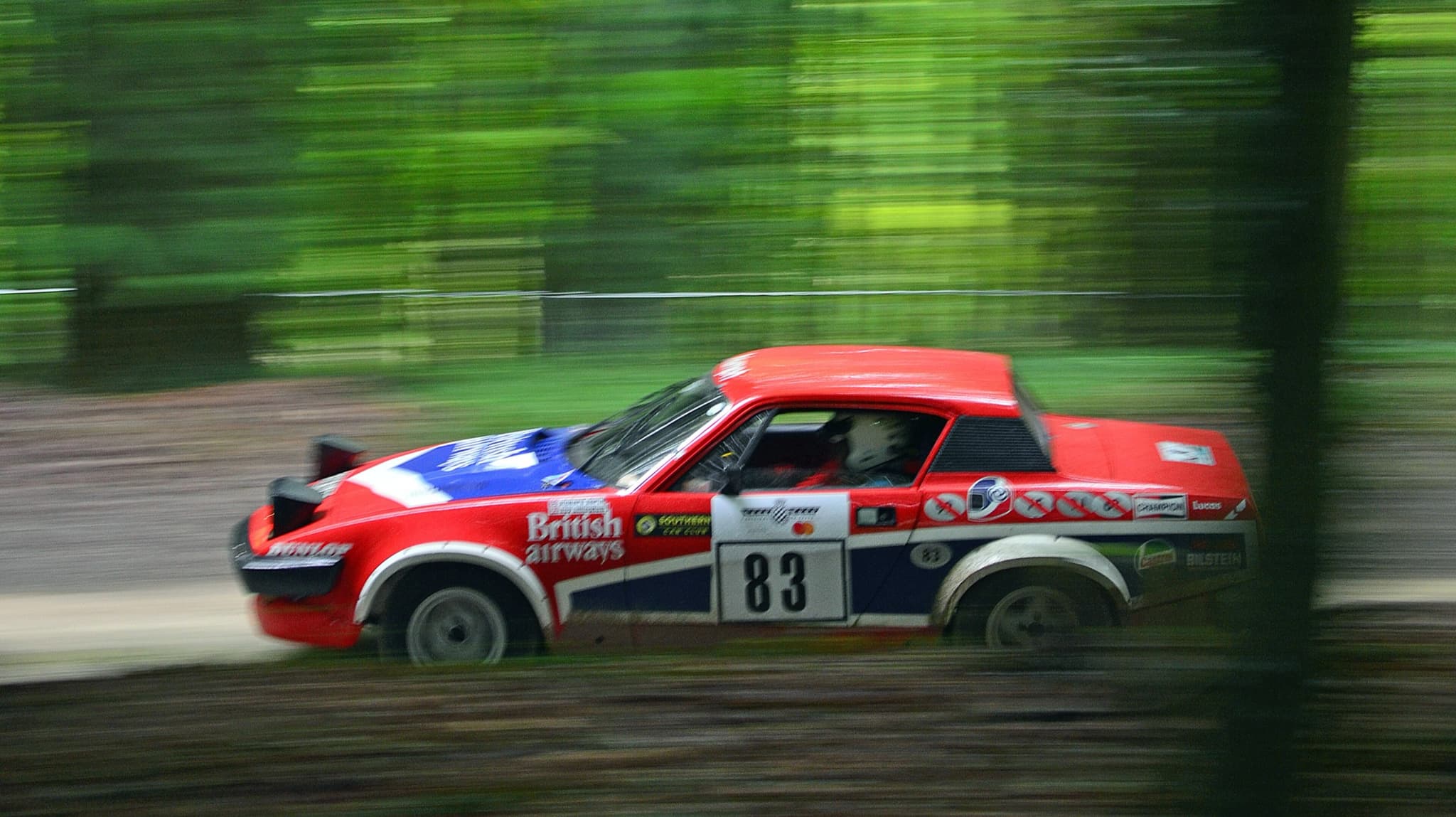 A rally coupé in red, white and blue airline colours, blurred trees behind as it speeds past.